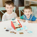 Two kids playing with a wooden cash register playset and toy money at a table