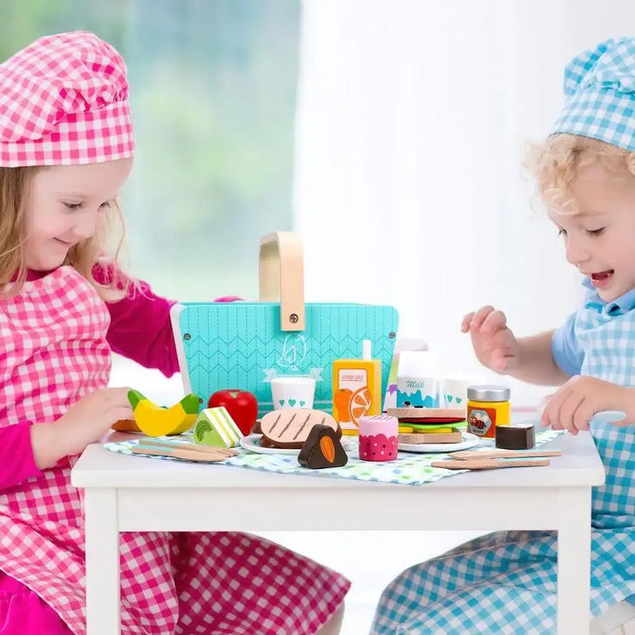 Kids in checkered aprons playing with wooden picnic basket playset and toy food on a table