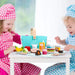 Kids in checkered aprons playing with wooden picnic basket playset and toy food on a table